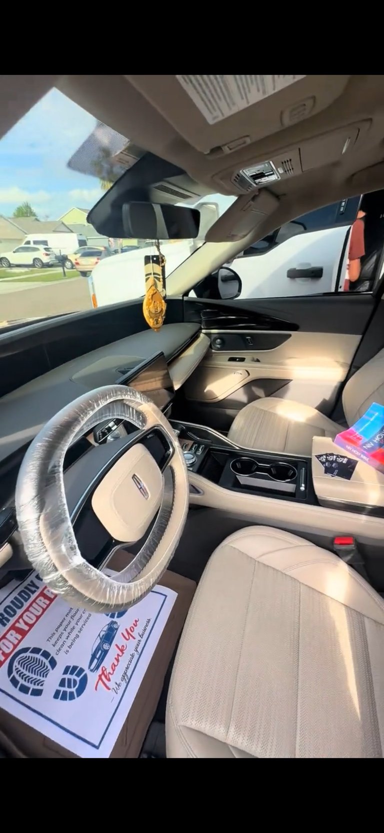 Interior of a new car on a dealership lot, showing tan and black upholstered seats, steering wheel wrapped in plastic, and protection film on surfaces