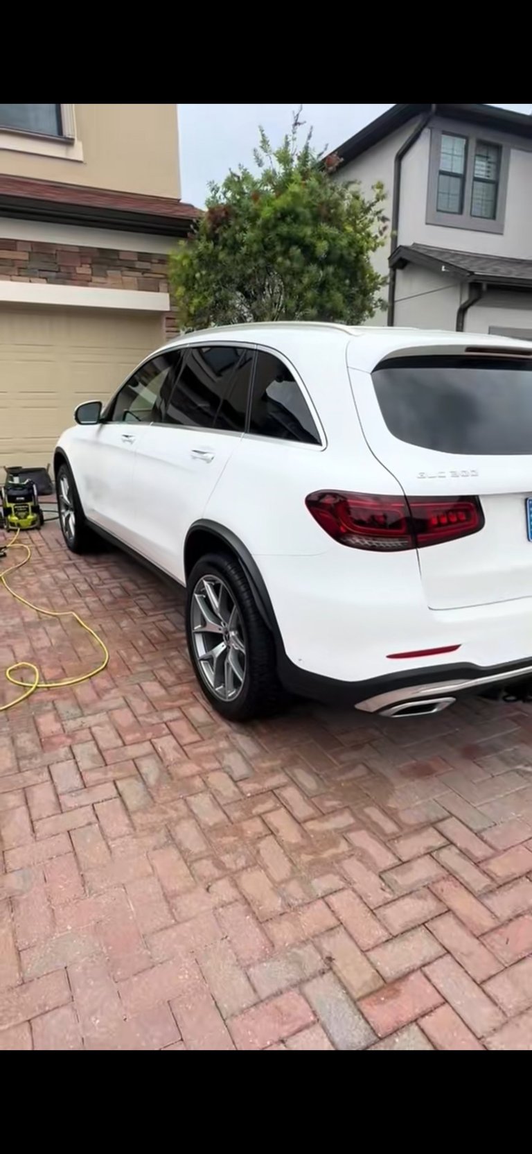 White Mercedes-Benz SUV parked in driveway with red brick pavement, garage and residential house visible in background