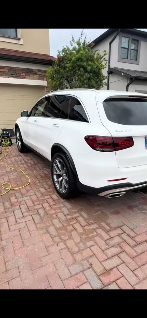 White Mercedes-Benz SUV parked in driveway with brick pavers, charging cable visible, residential house in background