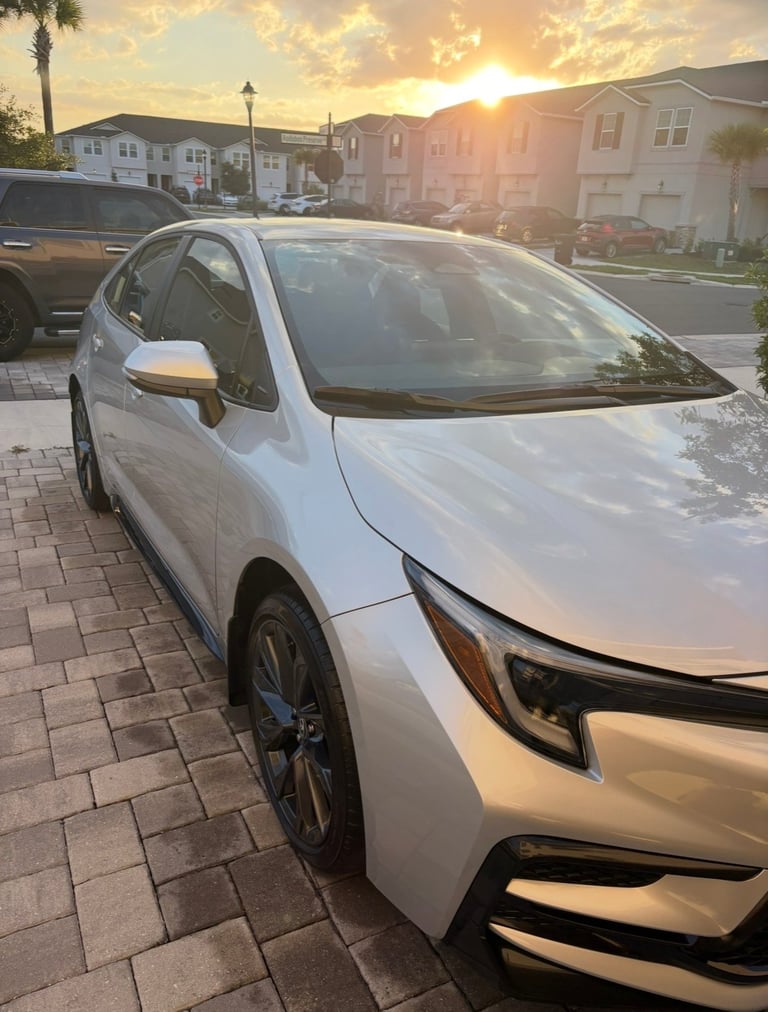 White compact car parked in residential driveway at sunset with houses and palm trees in background